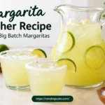 Margarita pitcher filled with fresh lime margaritas and ice, surrounded by two salt-rimmed margarita glasses, fresh limes, and a bowl of salt on a kitchen counter.